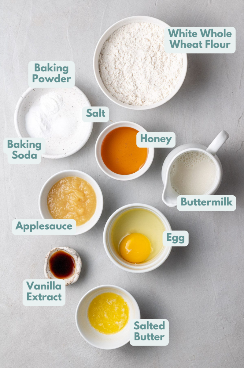 Healthy Pancake Recipe Ingredients in small white bowls on a grey background: white whole wheat flour, baking soda, baking powder, salt, salted butter, applesauce, egg, vanilla extract, honey or maple syrup, buttermilk