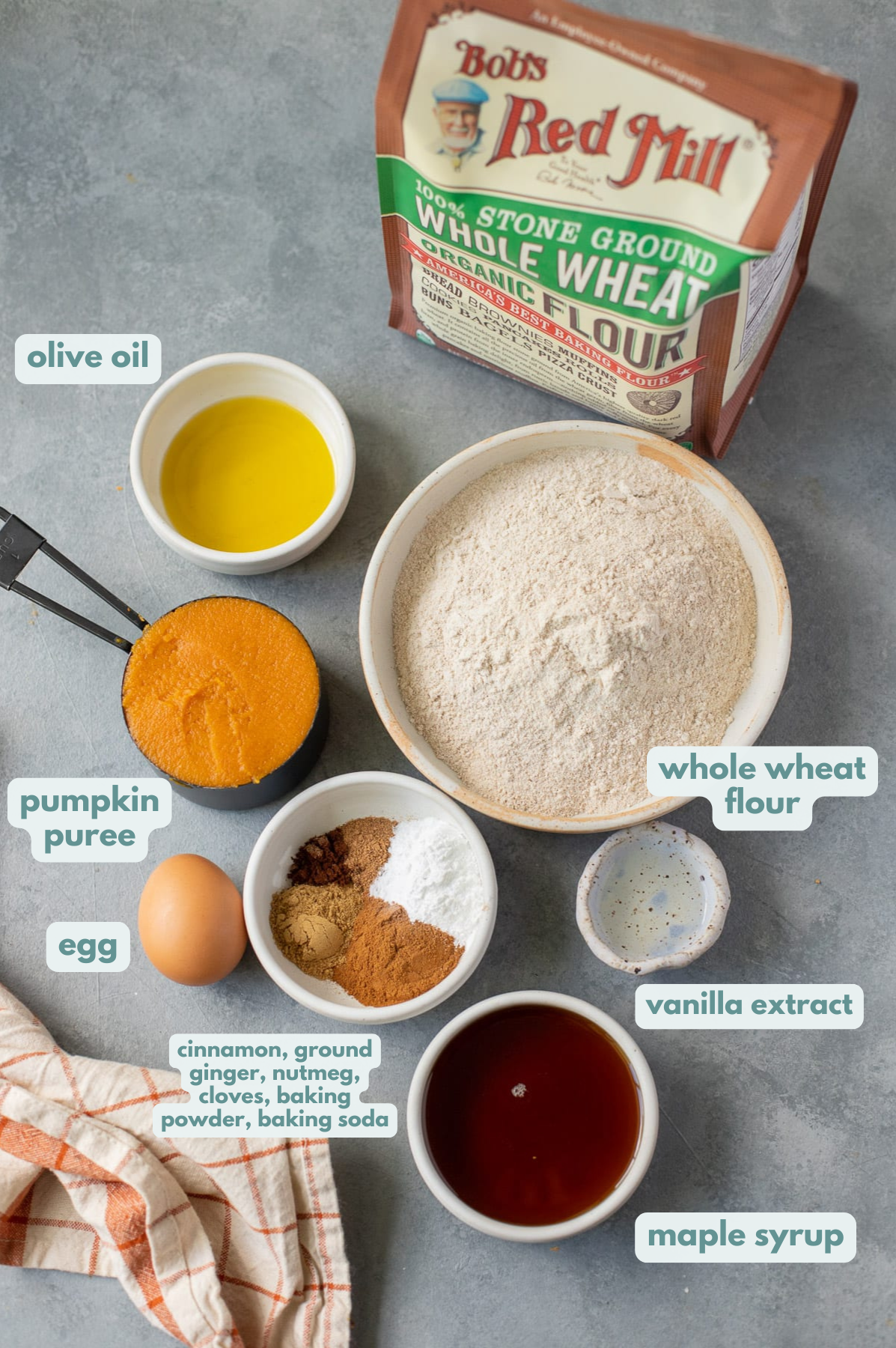 Healthy Pumpkin Bread Ingredients in small white bowls on a grey background: olive oil, pumpkin puree, egg, spices, baking powder, baking soda, maple syrup, vanilla extract, whole wheat flour.