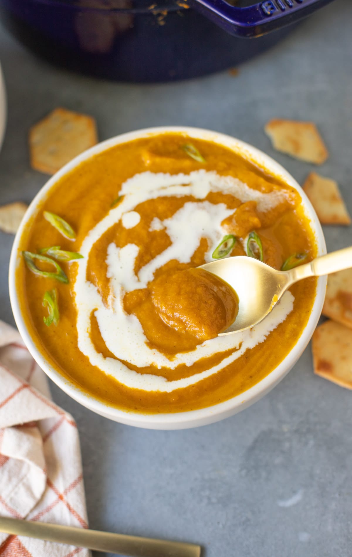 Spoonful of pumpkin soup being held above a bowl holding more of the creamy soup, garnished with a drizzle of heavy cream and sliced green onions.