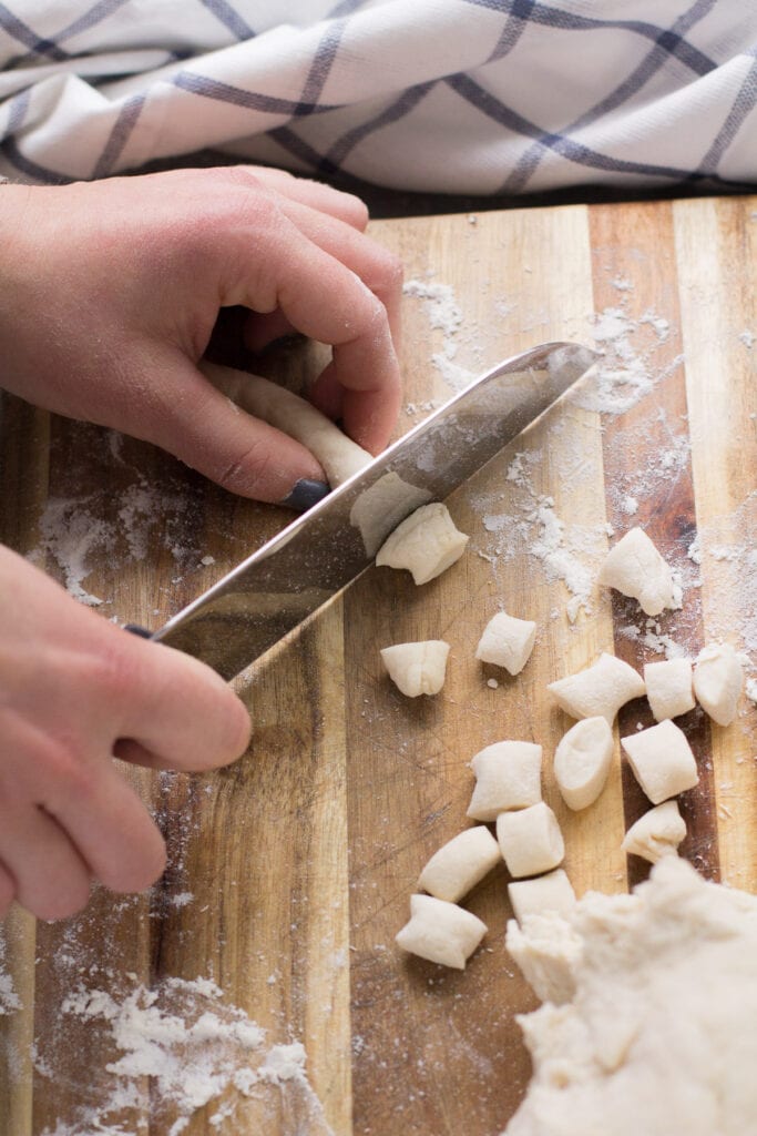 Hands using a knife to cut pieces of dough on a wooden cutting board.