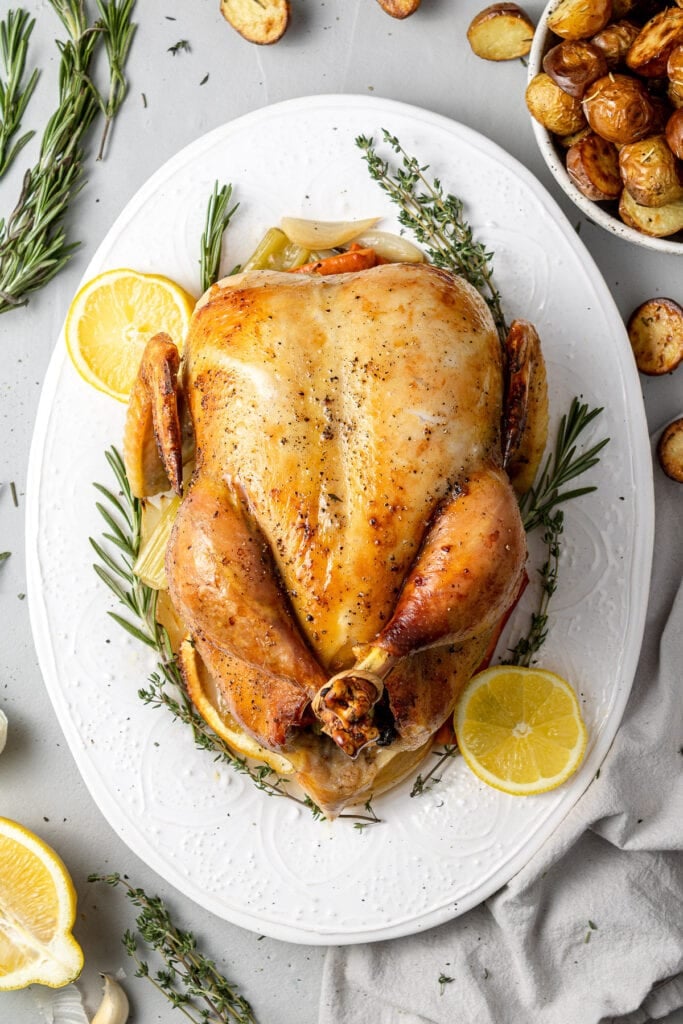 A close up of a juicy oven roasted chicken on a white platter decorated with fresh herbs