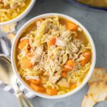 A bowl of turkey noodle soup on a grey background with a pot off to the side
