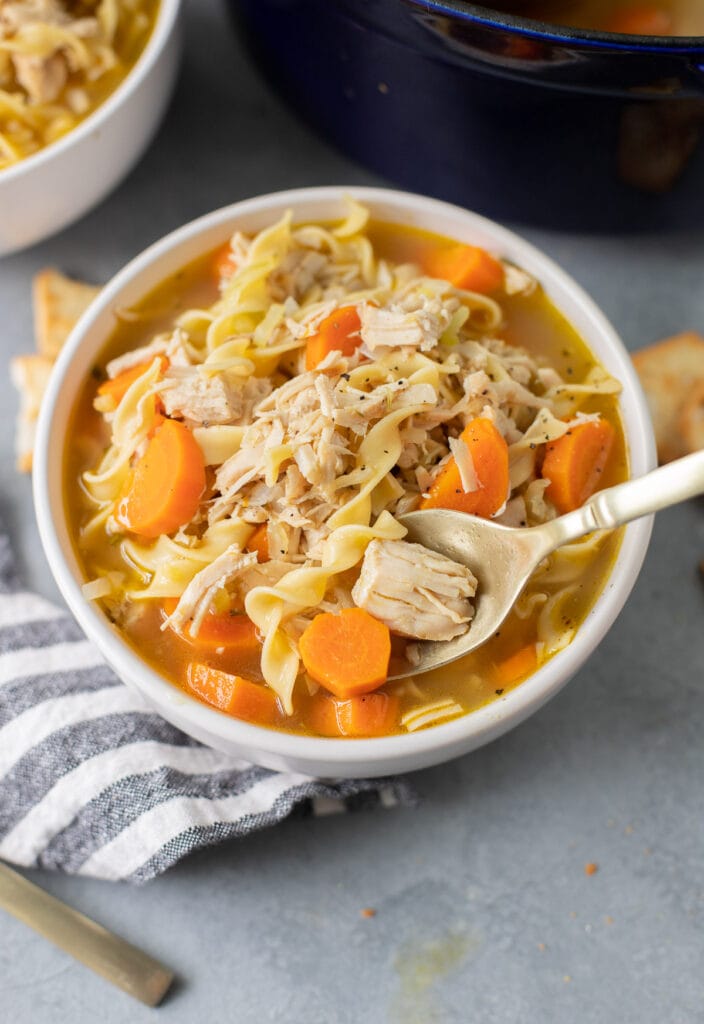 A close up of a bowl of turkey noodle soup on a grey background with a spoon taking a bite out of it