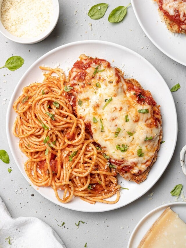 A plate of baked chicken parmesan with pasta and a bowl of cheese on the side