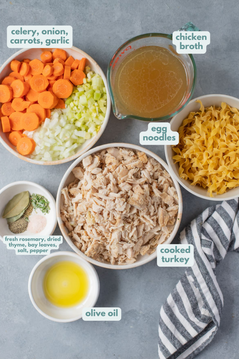 Leftover turkey noodle soup Ingredients in white bowls on a grey background: cooked turkey, celery, onion, carrots, turkey or chicken broth, egg noodles, olive oil, garlic, rosemary, thyme, red pepper flakes, bay leaves, salt & pepper