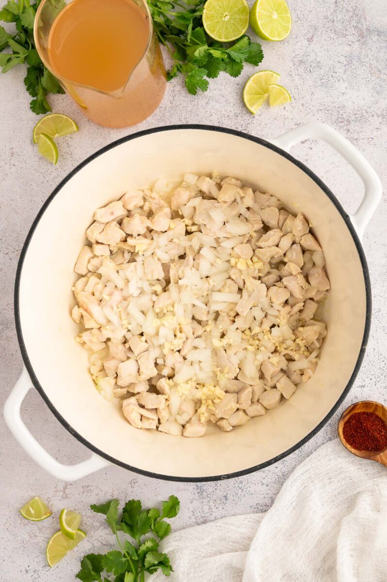 A dutch oven with cubed cooked chicken, garlic, and diced onion in it on a grey background.