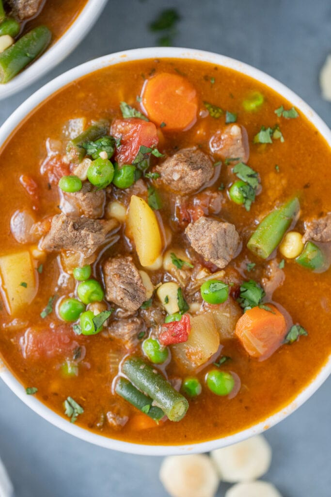 A close up of vegetable beef soup in a white bowl with a grey background