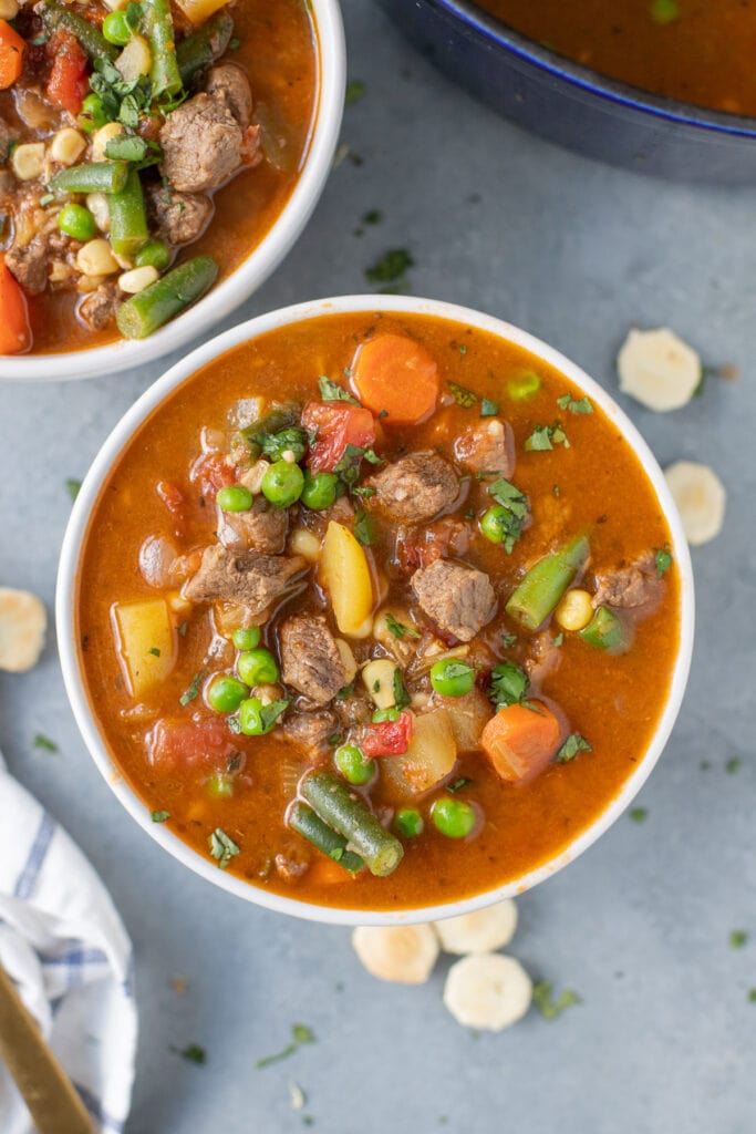 A white bowl of beef vegetable soup with another bowl on the side on a grey background