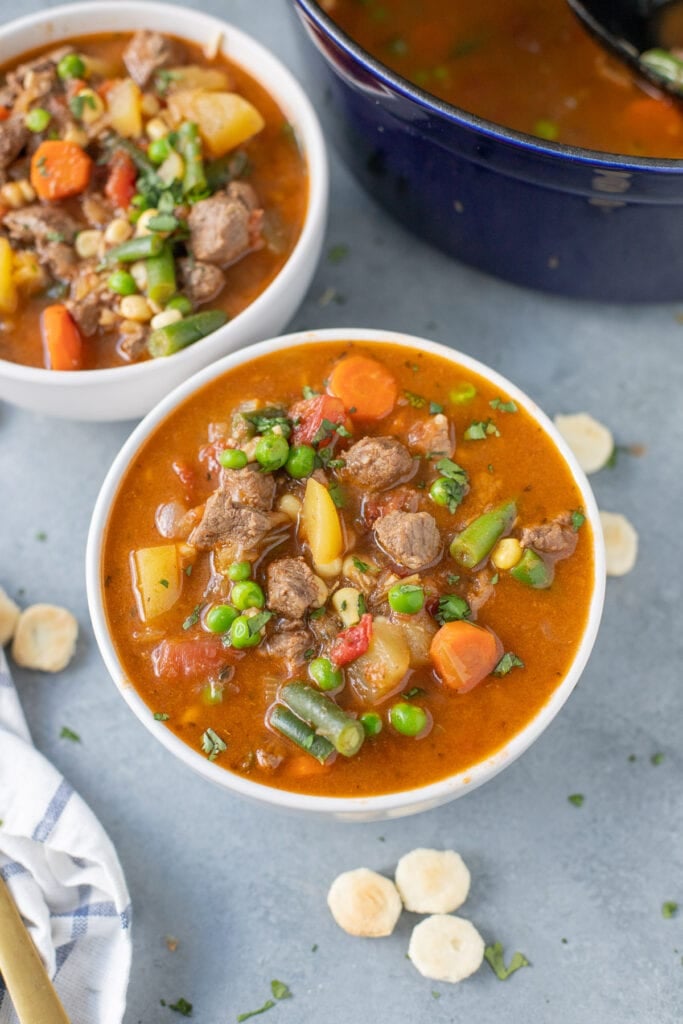 A white bowl of beef vegetable soup with another bowl on the side on a grey background