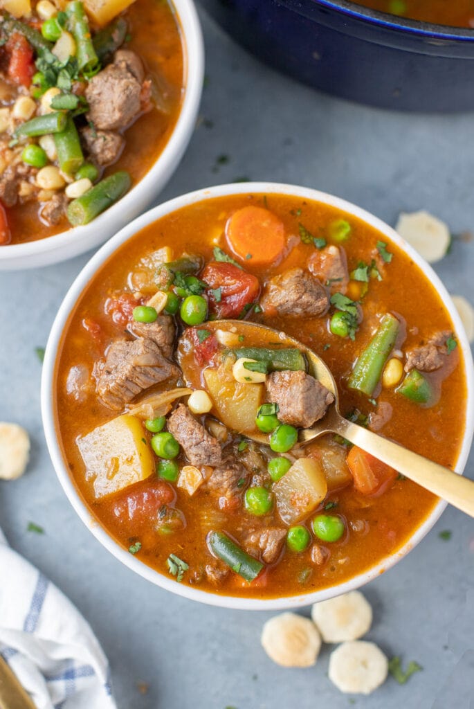 A spoon taking a bite out of a bowl of beef and vegetable soup in a white bowl on a grey background