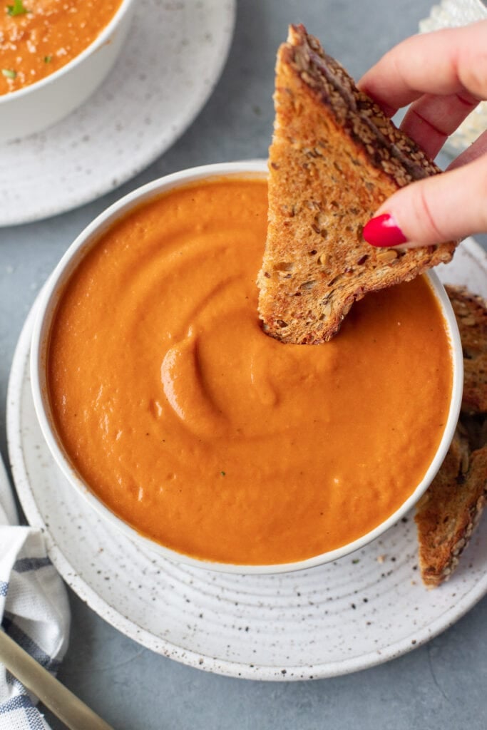 A bowl of creamy tomato basil soup with half of a grilled cheese being dipped in it