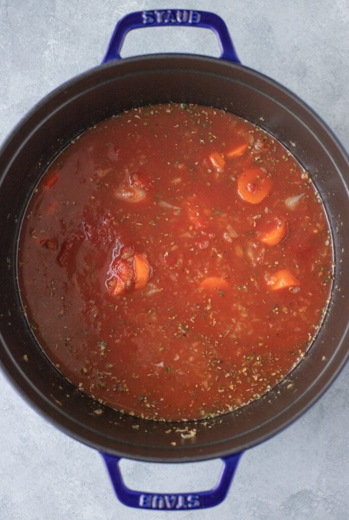 A stock pot full of crushed tomatoes, broth, dried spices, sauteed carrots, onions and garlic for tomato basil soup