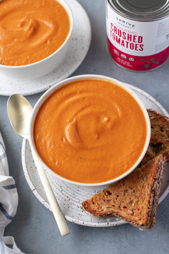 A bowl of tomato basil soup with another bowl on the side and a grilled cheese on the side
