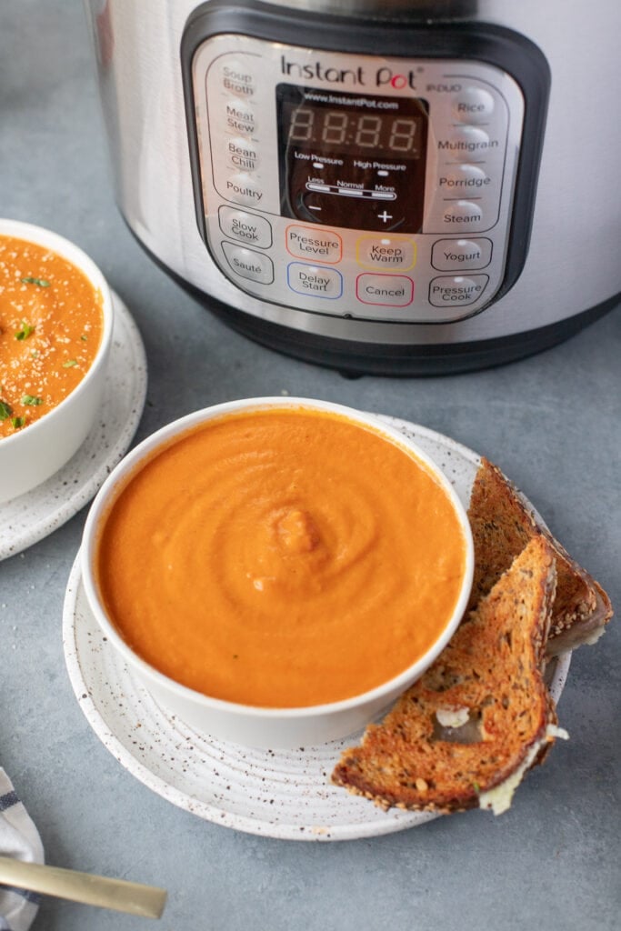 A bowl of tomato basil soup and a grilled cheese on the side with an instant pot behind it