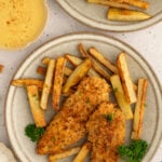 Baked chicken tenders + fries on a plate with a another plate of food and a bowl of honey mustard off to the side.