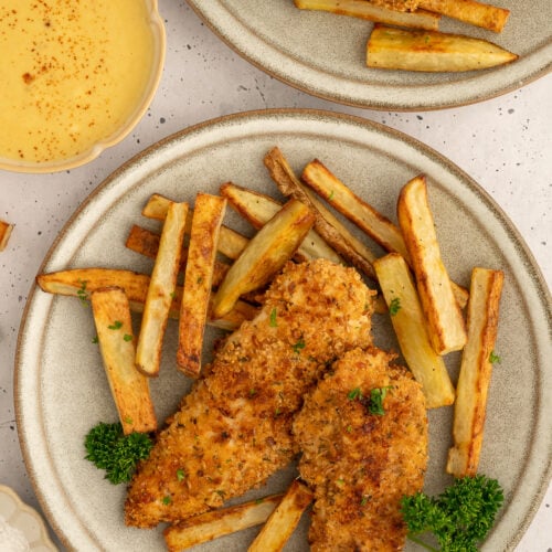 Baked chicken tenders + fries on a plate with a another plate of food and a bowl of honey mustard off to the side.