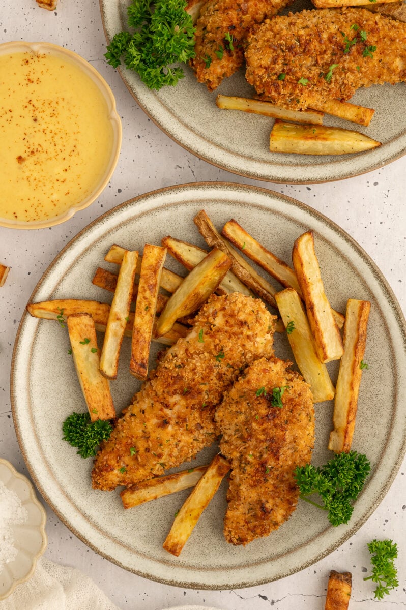 Baked chicken tenders + fries on a plate with a another plate of food and a bowl of honey mustard off to the side.