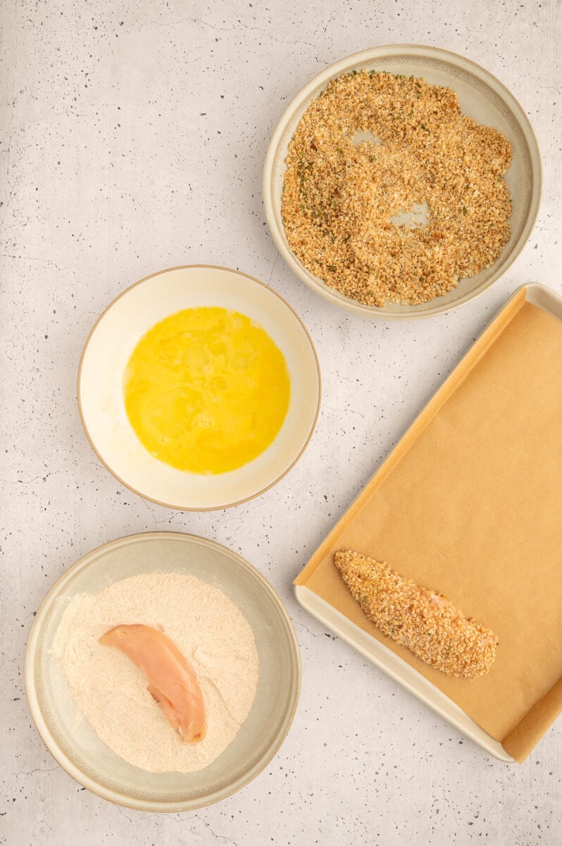 A raw chicken tender being dipped in flour mixture to make healthy chicken tenders with two other bowls of beaten egg and breadcrumbs and a breaded chicken tender on a parchment lined baking sheet.