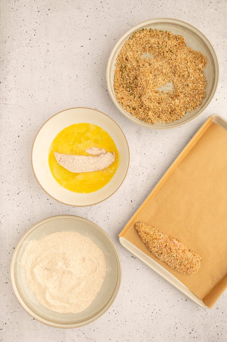 A raw chicken tender being dipped in egg mixture to make healthy chicken tenders with other bowls of flour and breadcrumbs on the side.