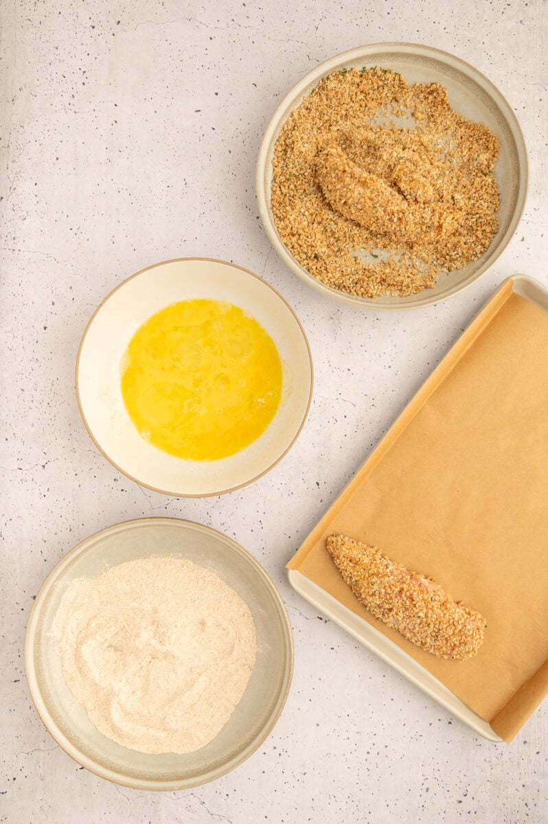 A raw chicken tender being dipped in breadcrumb mixture to make healthy chicken tenders with other bowls of flour and beaten egg on the side and a breaded chicken tender on a parchment lined baking sheet.