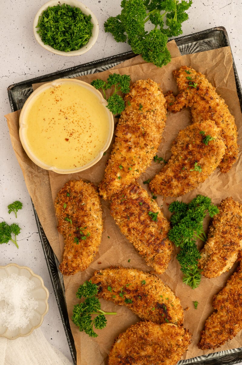Baked Chicken Tenders on a baking sheet with fresh herbs and a bowl of honey mustard.