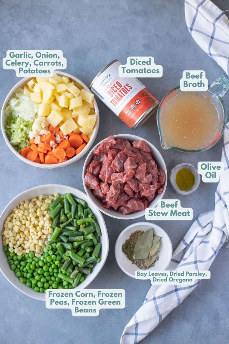 Ingredients for vegetable beef soup in white bowls: beef stew meat, olive oil, onions, celery, carrots, potatoes, garlic, beef broth, petite diced tomatoes, bay leaves, dried parsley, dried basil, dried oregano, frozen corn, frozen peas, frozen green beans
