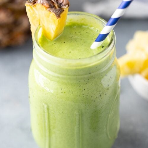 A side shot of a green smoothie in a mason jar with a pineapple on the rim with a blue and white straw in it