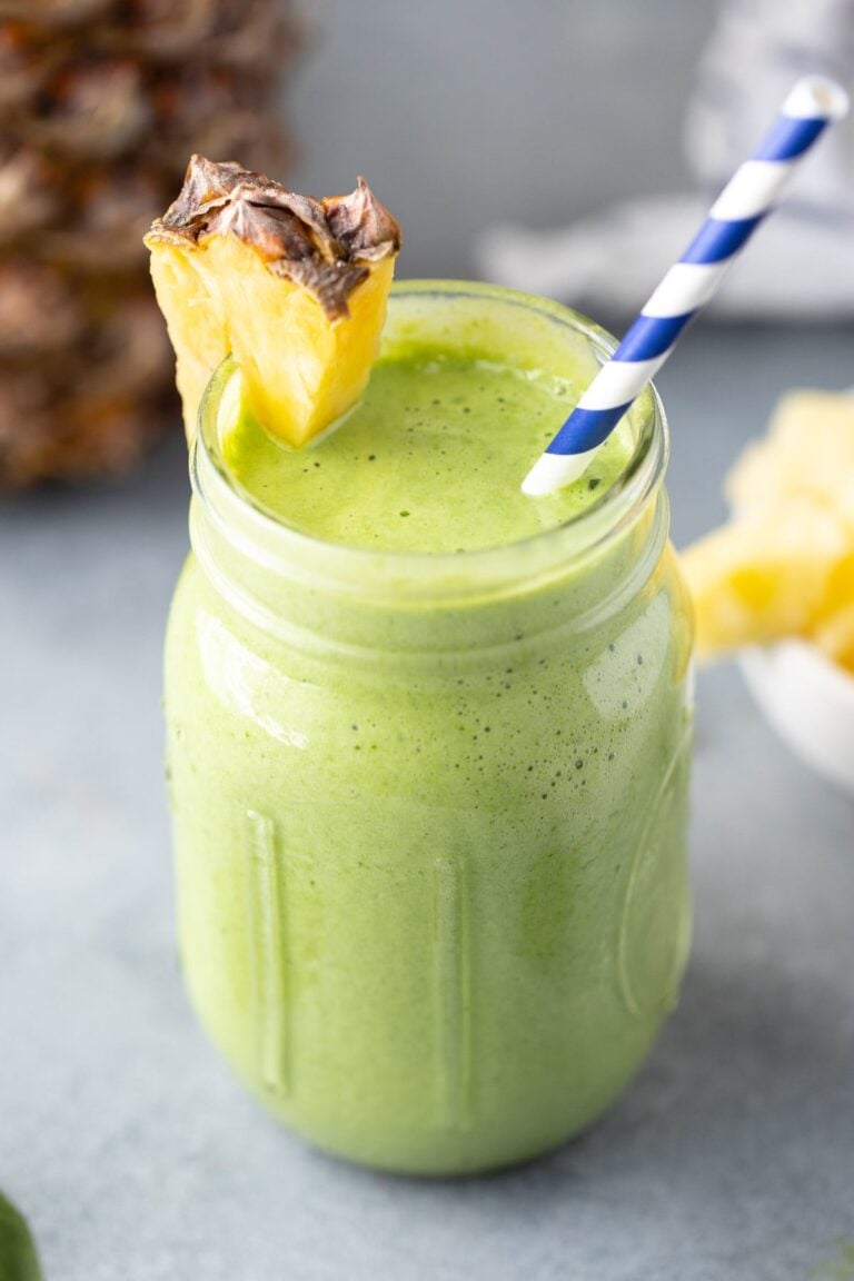 A side shot of a green smoothie in a mason jar with a pineapple on the rim with a blue and white straw in it