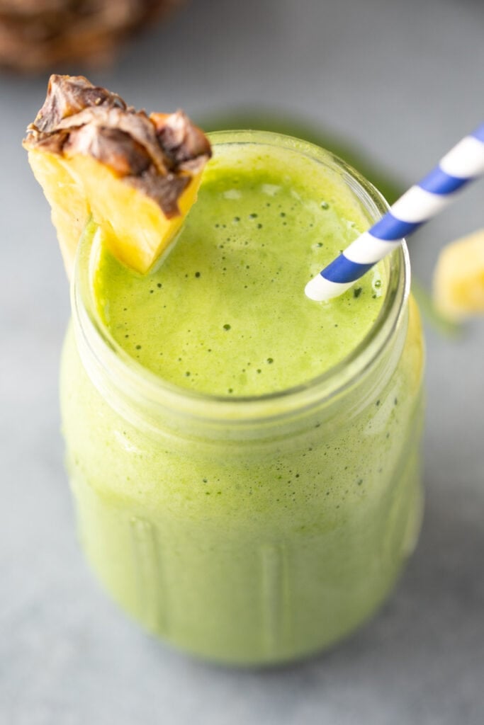 A green smoothie in a mason jar with a pineapple on the rim with a blue and white straw in it