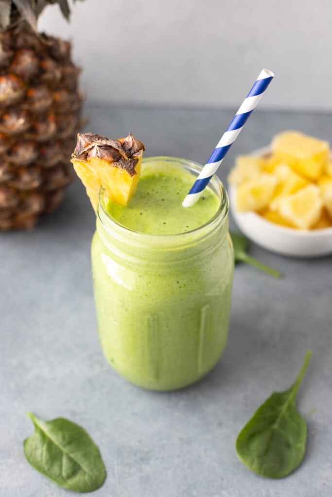 A side shot of a green smoothie in a mason jar with a pineapple on the rim with a blue and white straw in it with pineapple and spinach on the side