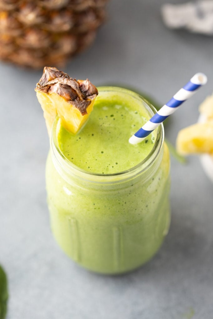 A green smoothie in a mason jar with a pineapple on the rim with a blue and white straw in it