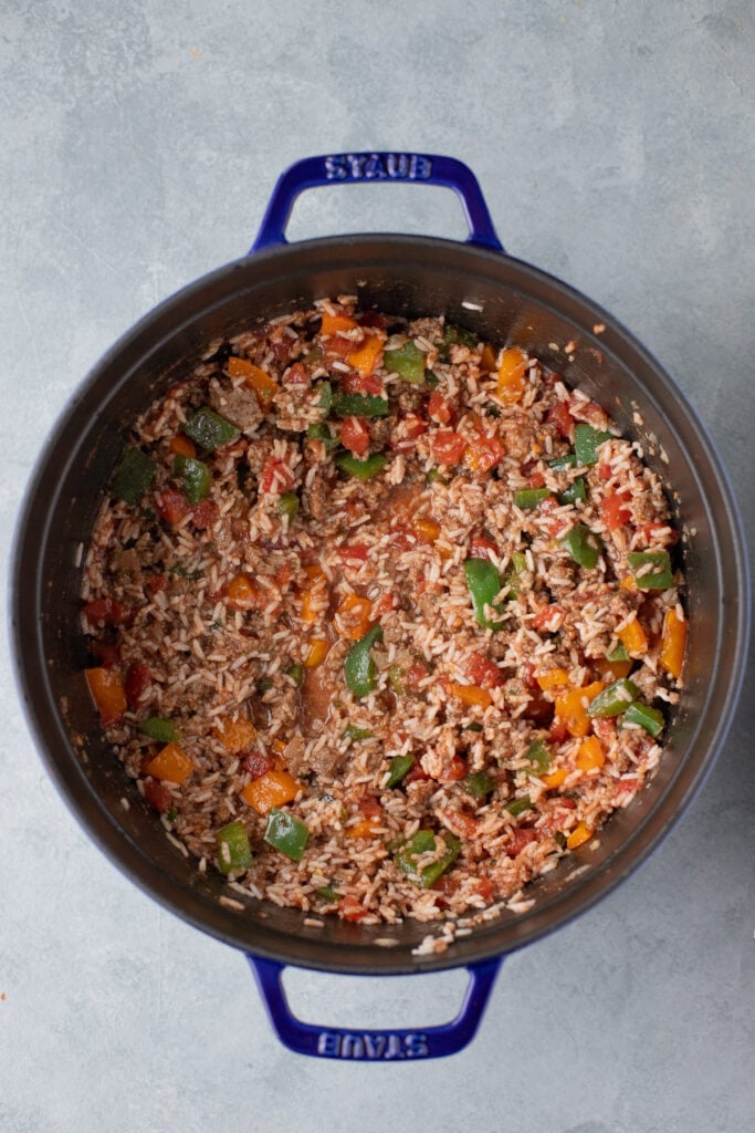 a dutch oven with garlic, onion, peppers, beef, tomato sauce, tomatoes, salt, pepper, oregano, basil, rice and beef broth mixed for stuffed bell pepper casserole
