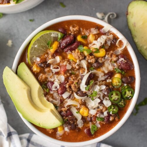 A bowl of taco soup with avocado and lime in the bowl on a grey background