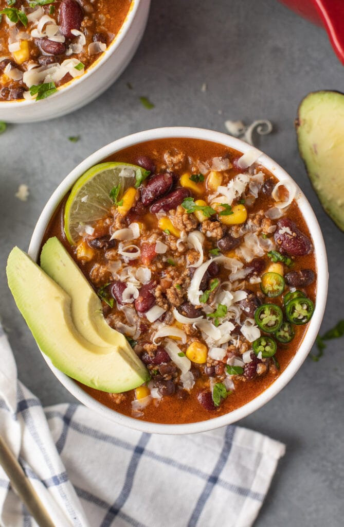 A bowl of taco soup with avocado and lime in the bowl on a grey background