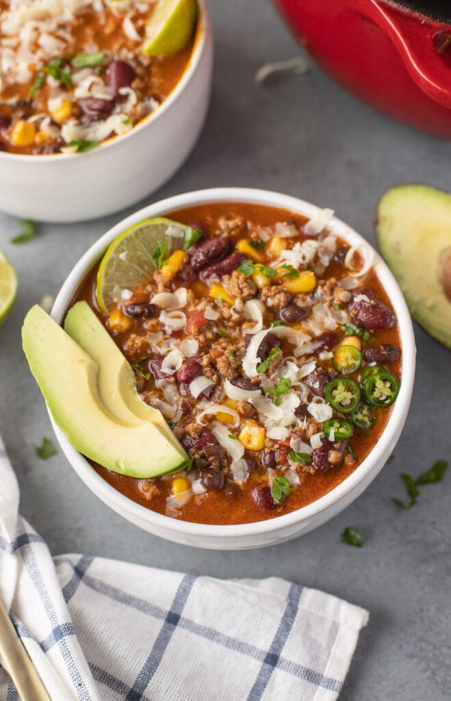 A bowl of taco soup with avocado and lime in the bowl on a grey background with another bowl of soup on the side