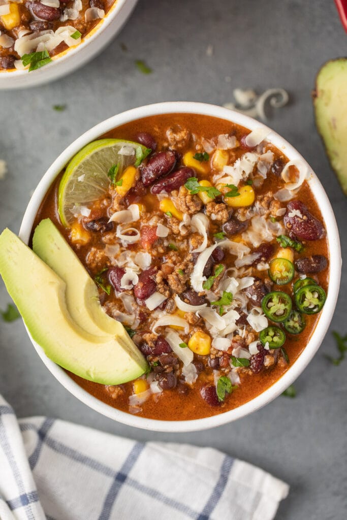 A close up bowl of taco soup with avocado and lime in the bowl on a grey background