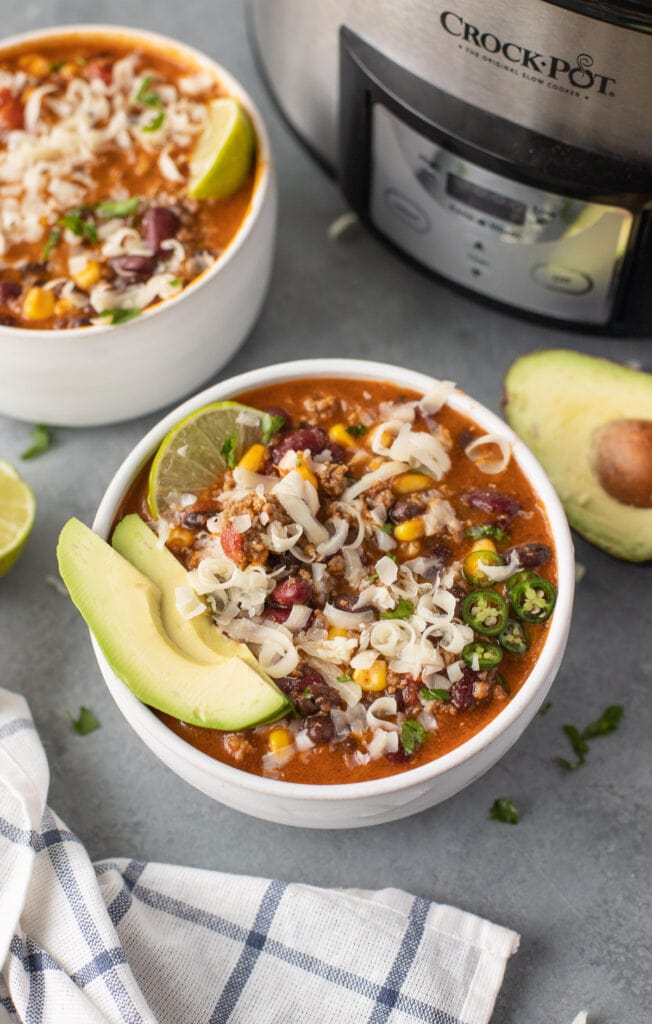 A bowl of taco soup with avocado and lime in the bowl on a grey background with another bowl of soup and crock pot on the side