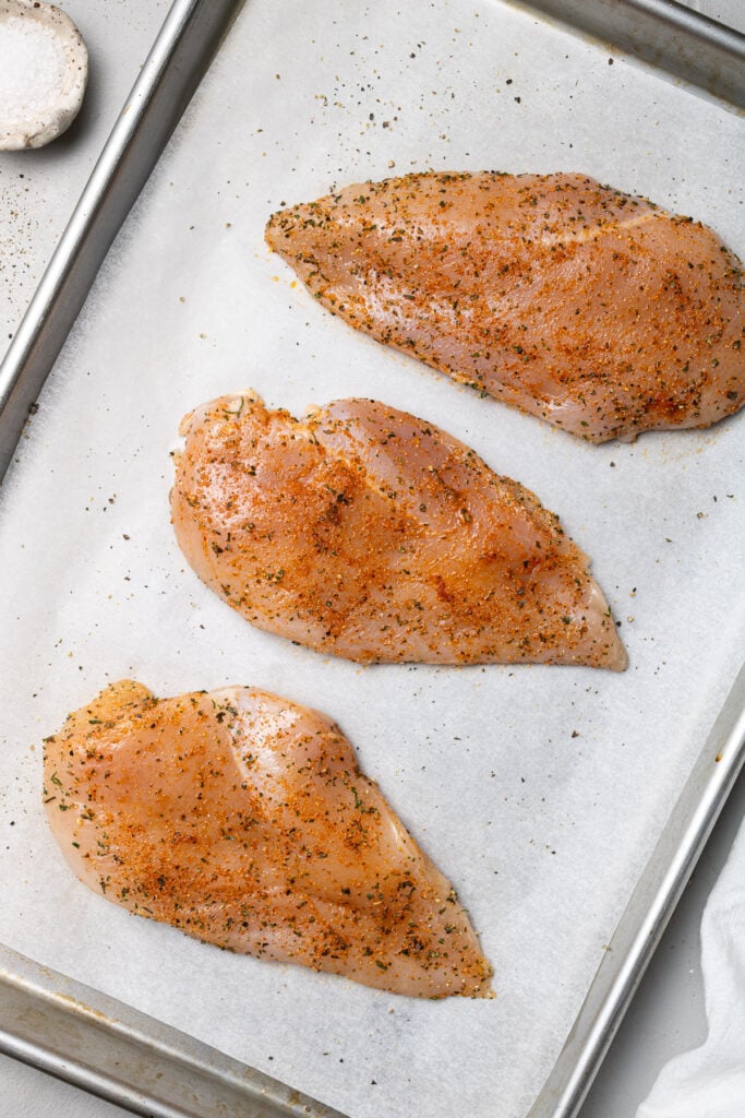 Uncooked boneless chicken breasts covered in olive oil and spices on a parchment lined pan