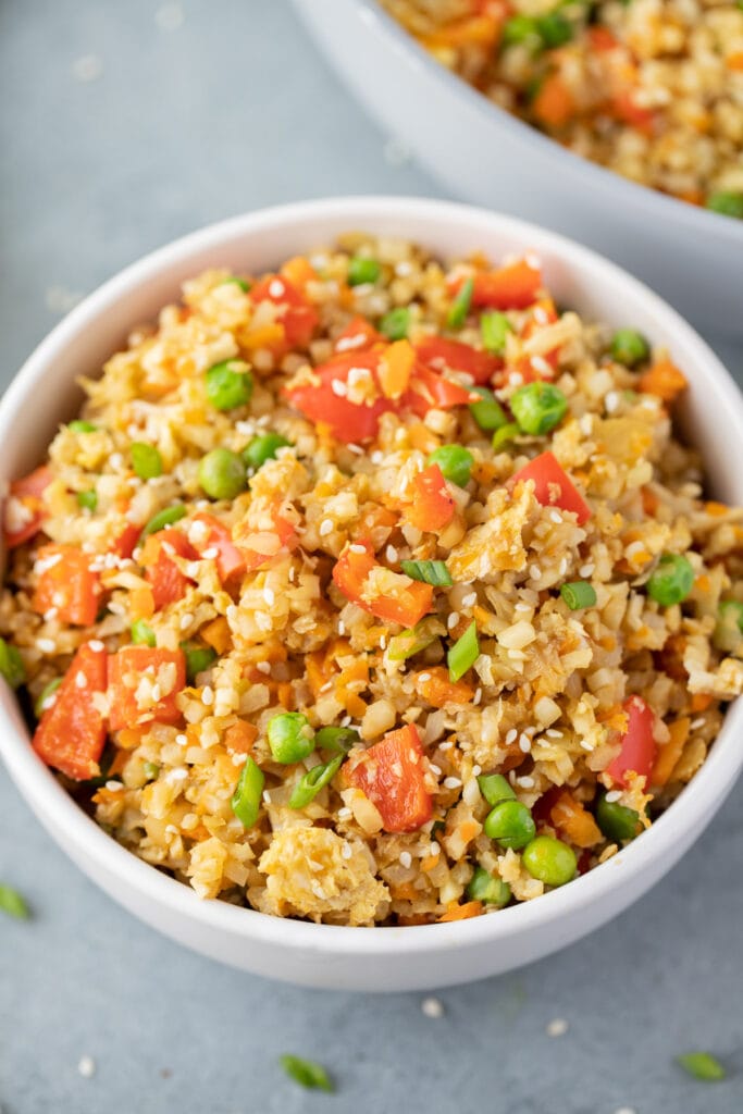 An overhead shot of fried cauliflower rice in a white bowl on a grey background with another bowl on the side