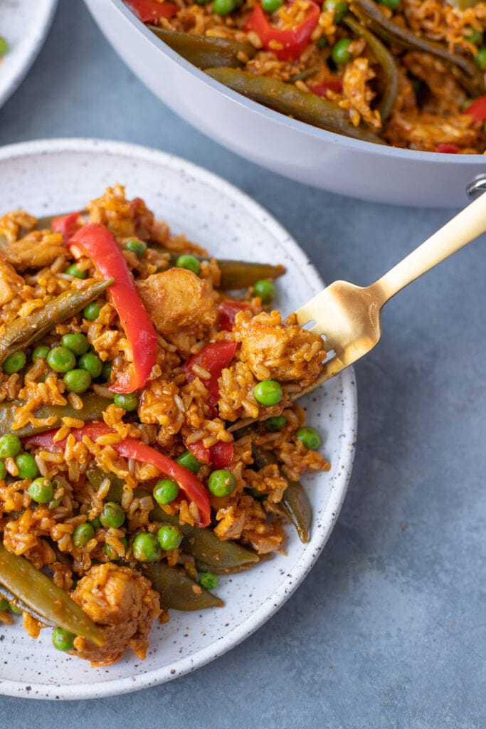 An overhead shot of chicken paella on a white plate with a bite being taken out of it on a grey background