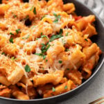 A black bowl with chicken parmesan pasta with fresh herbs and cheese on top with gold spoons on the side