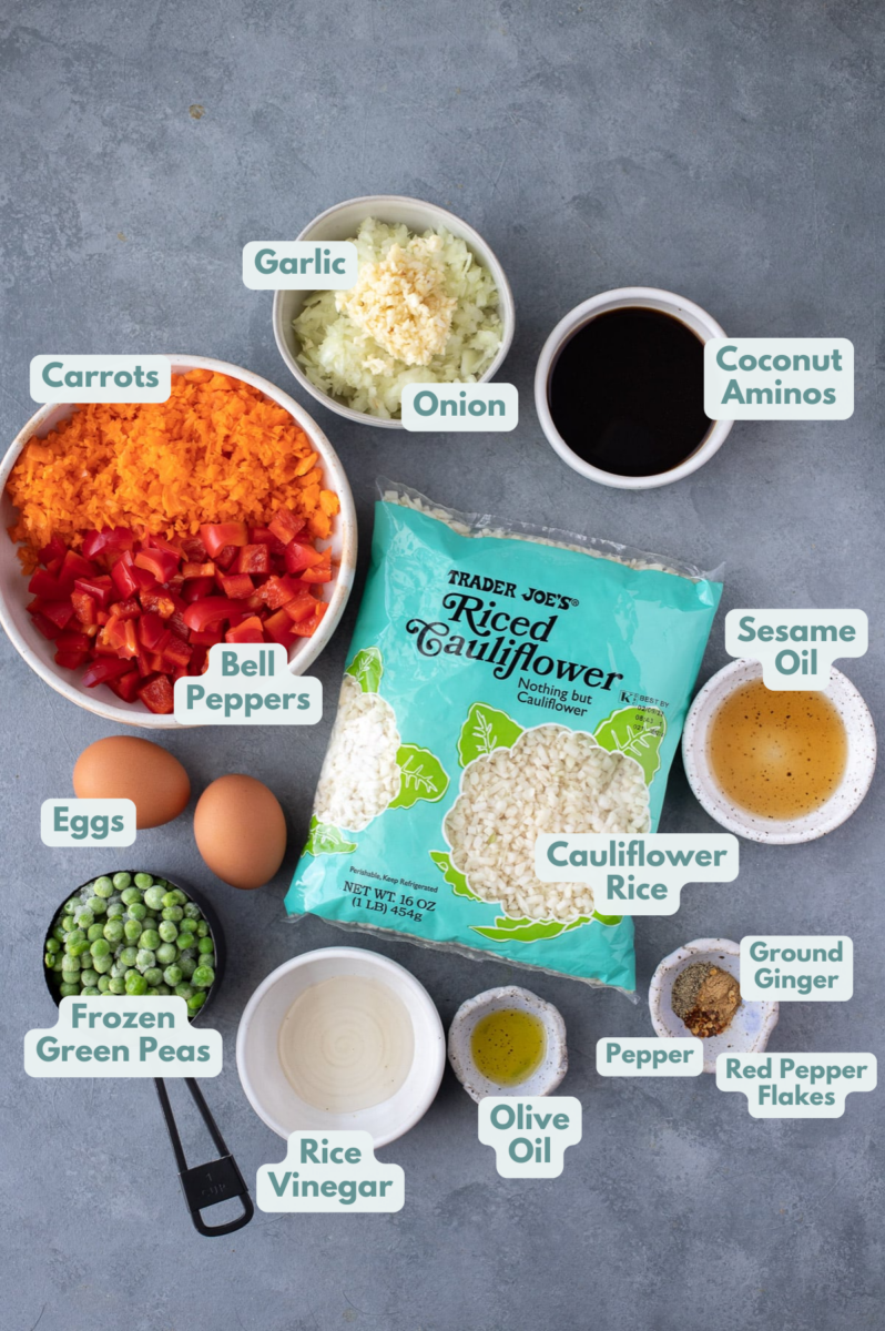 Ingredients for fried cauliflower rice in white bowls: olive oil, onion, garlic, red bell pepper, carrots, cauliflower rice, frozen green peas, eggs, ground ginger, red pepper flakes, pepper, toasted sesame oil, rice wine vinegar, coconut aminos