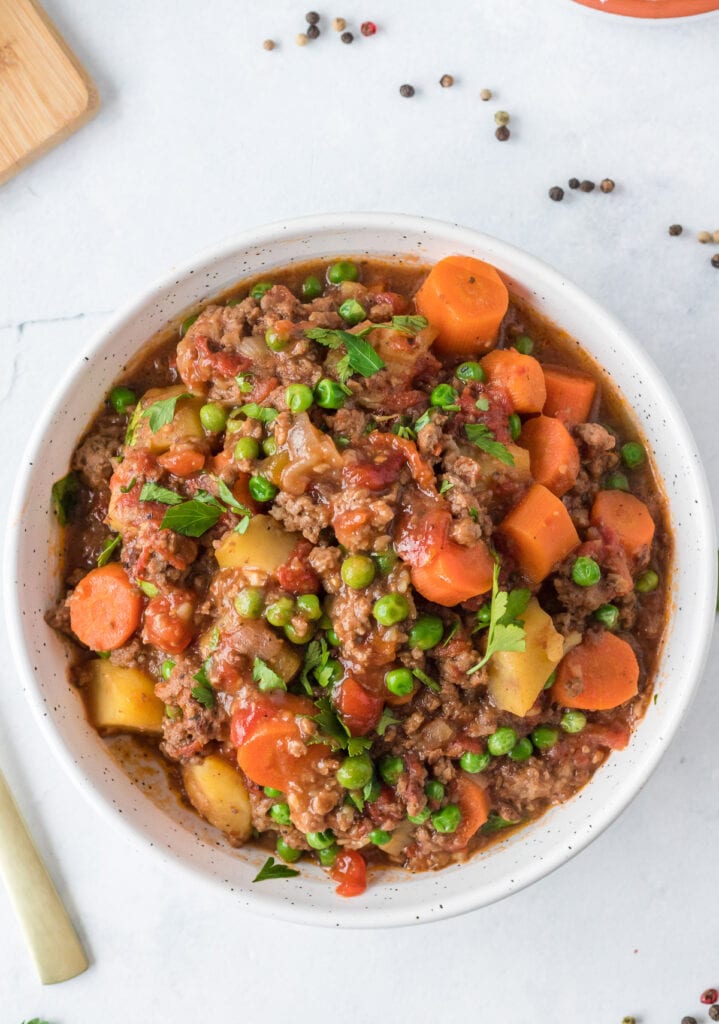 A close up of a bowl of ground beef stew on a white background with a spoon and ingredients off to the side