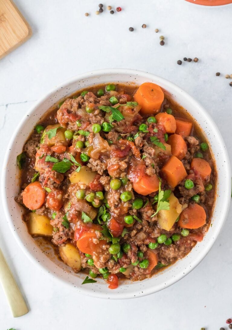 A close up of a bowl of ground beef stew on a white background with a spoon and ingredients off to the side