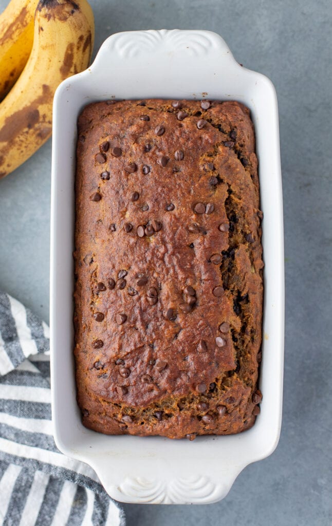 a freshly cooked healthy banana bread in a white loaf pan with bananas on the side