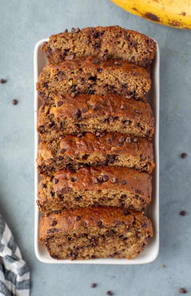 Healthy Banana Bread cut into slices on a blue background with bananas, chocolate chips + linen off to the side