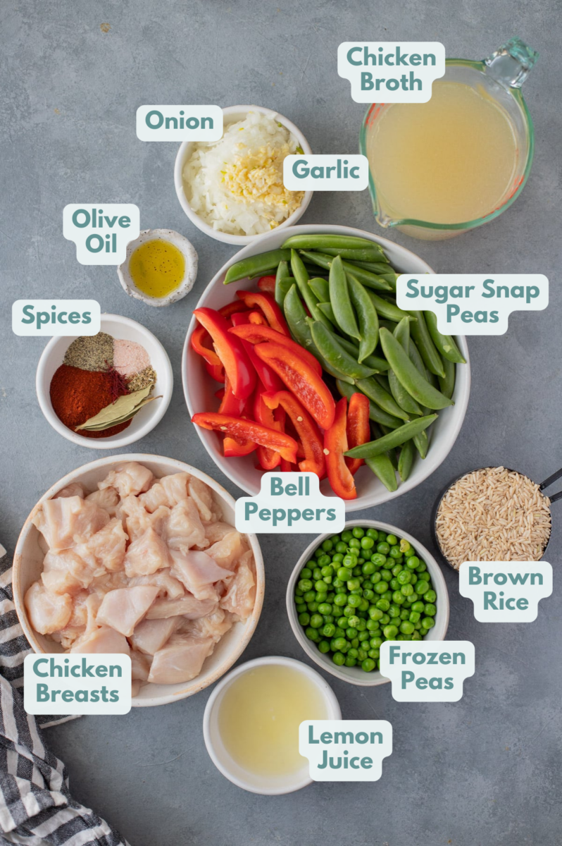 Chicken paella ingredients in white bowls: chicken breasts, olive oil, onion, garlic, bell peppers, sugar snap peas, pepper, salt, paprika, bay leaves, dried thyme, saffron, lemon juice, brown rice, chicken broth, frozen peas