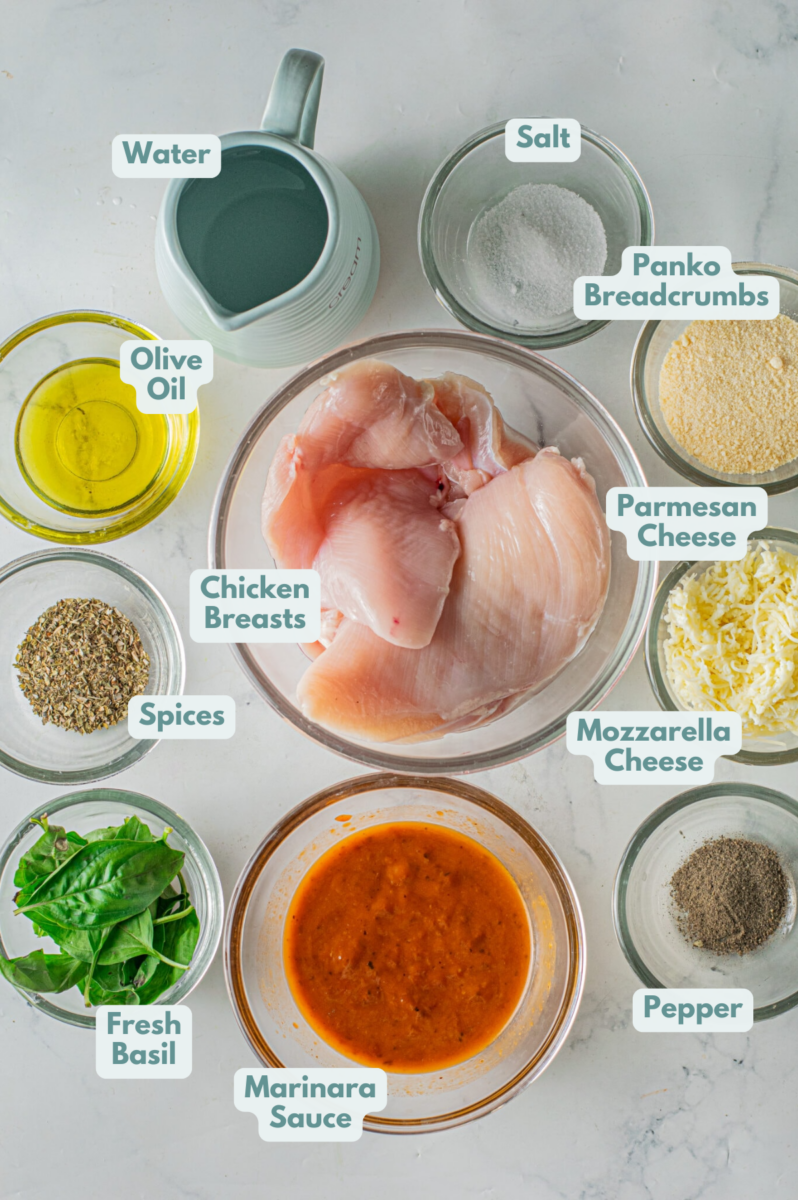Ingredients for instant pot chicken parmesan in clear bowls on a grey background: boneless, skinless chicken breasts, olive oil, marinara sauce, water, salt, pepper dried parsley, dried basil, dried oregano, garlic powder, fresh basil, shredded mozzarella cheese, grated parmesan cheese