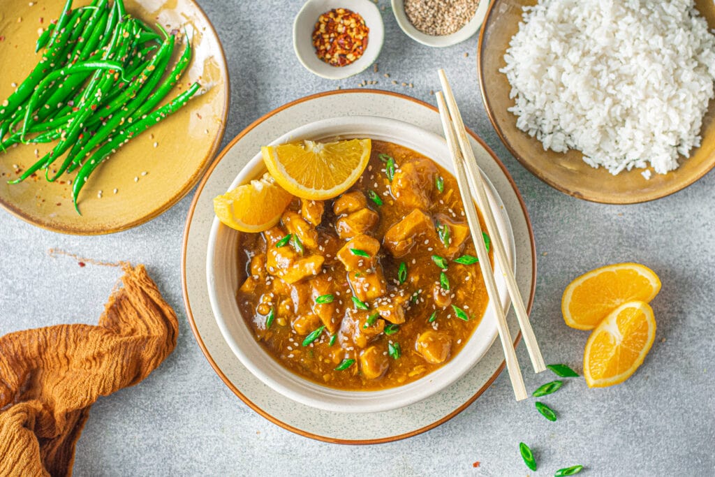 a horizontal image of the best easy orange chicken on a grey background with garnishes off to the side