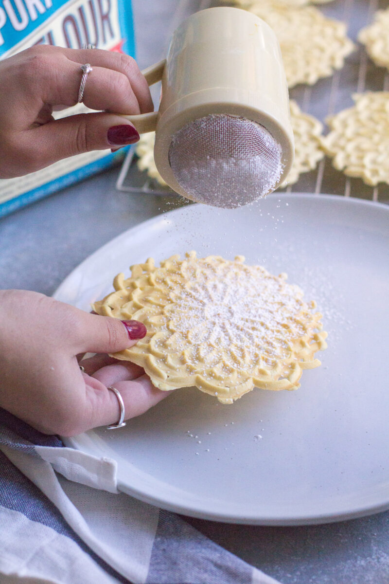 A hands shaking powdered sugar over a pizzelle cookie.